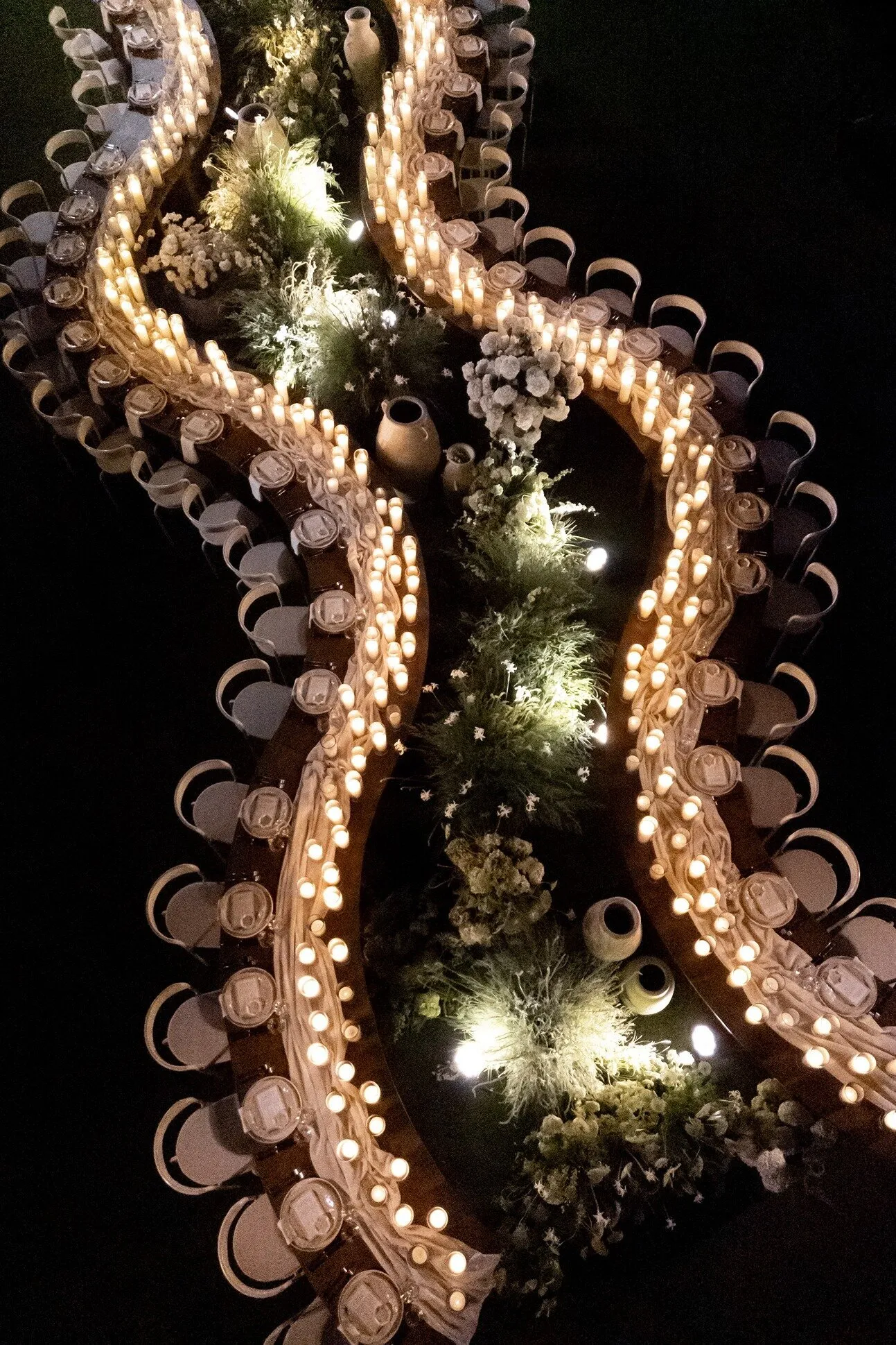 A serpentine dinner table seen from above at night, lit entirely by candles