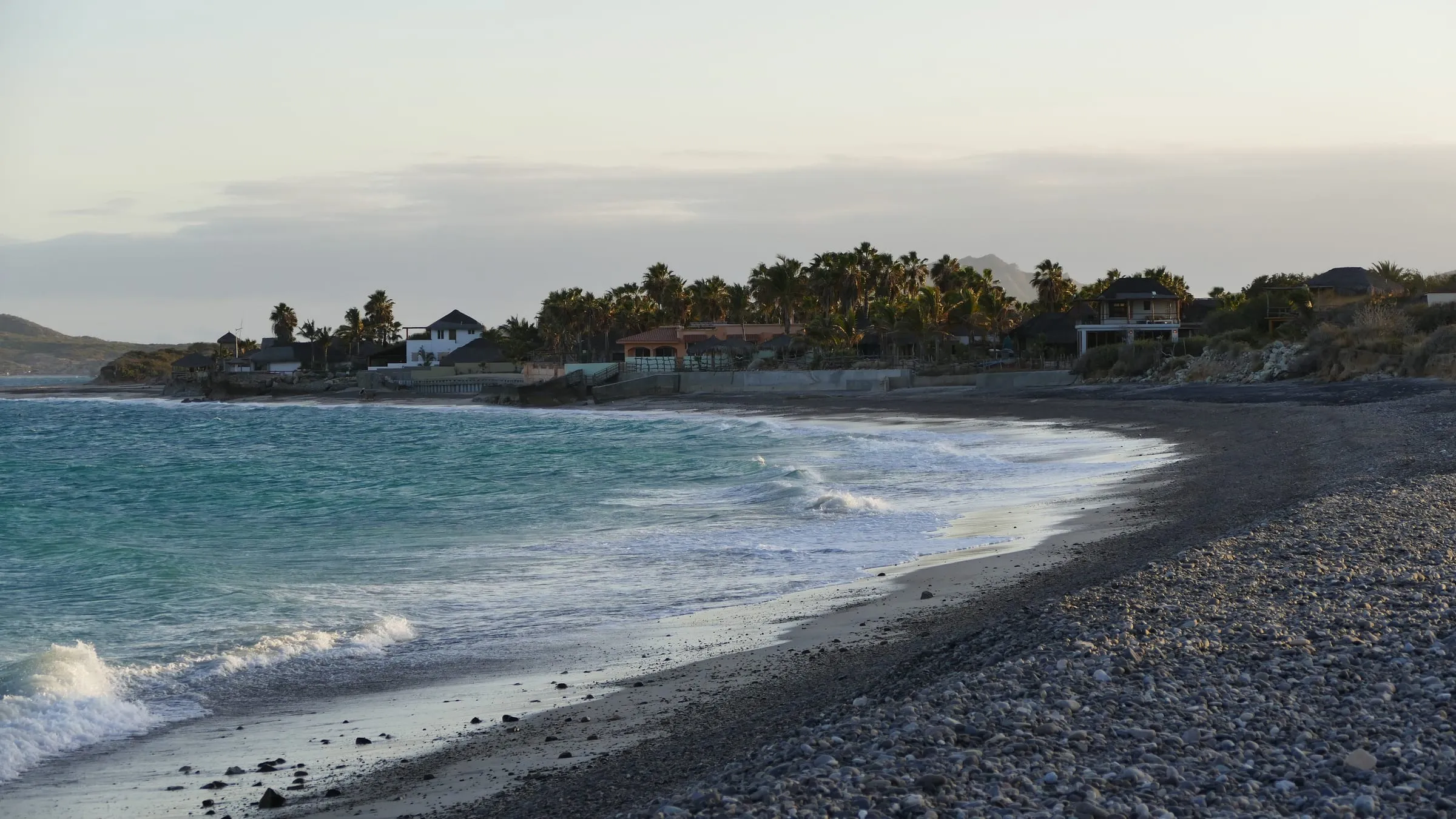 A curved pebble beach at dusk on the East Cape, low houses among the palms