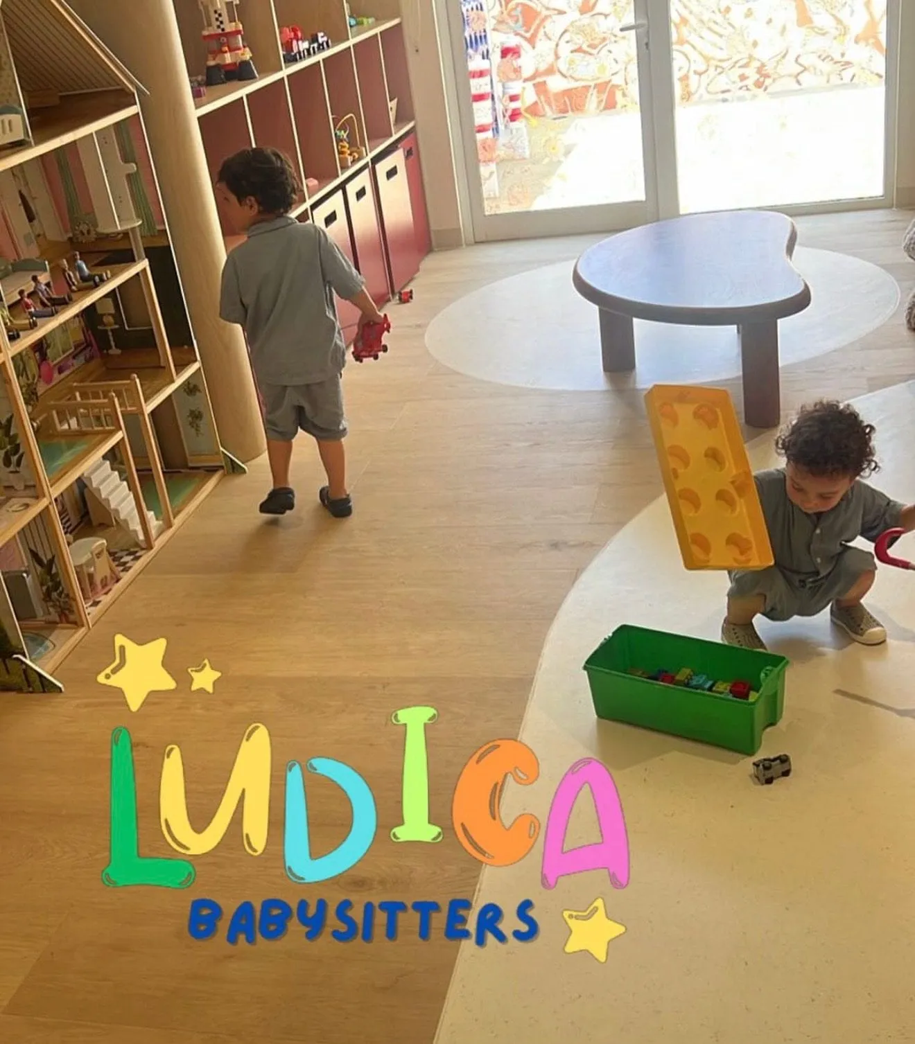 Two small children in a playroom, one carrying a foam block across the floor