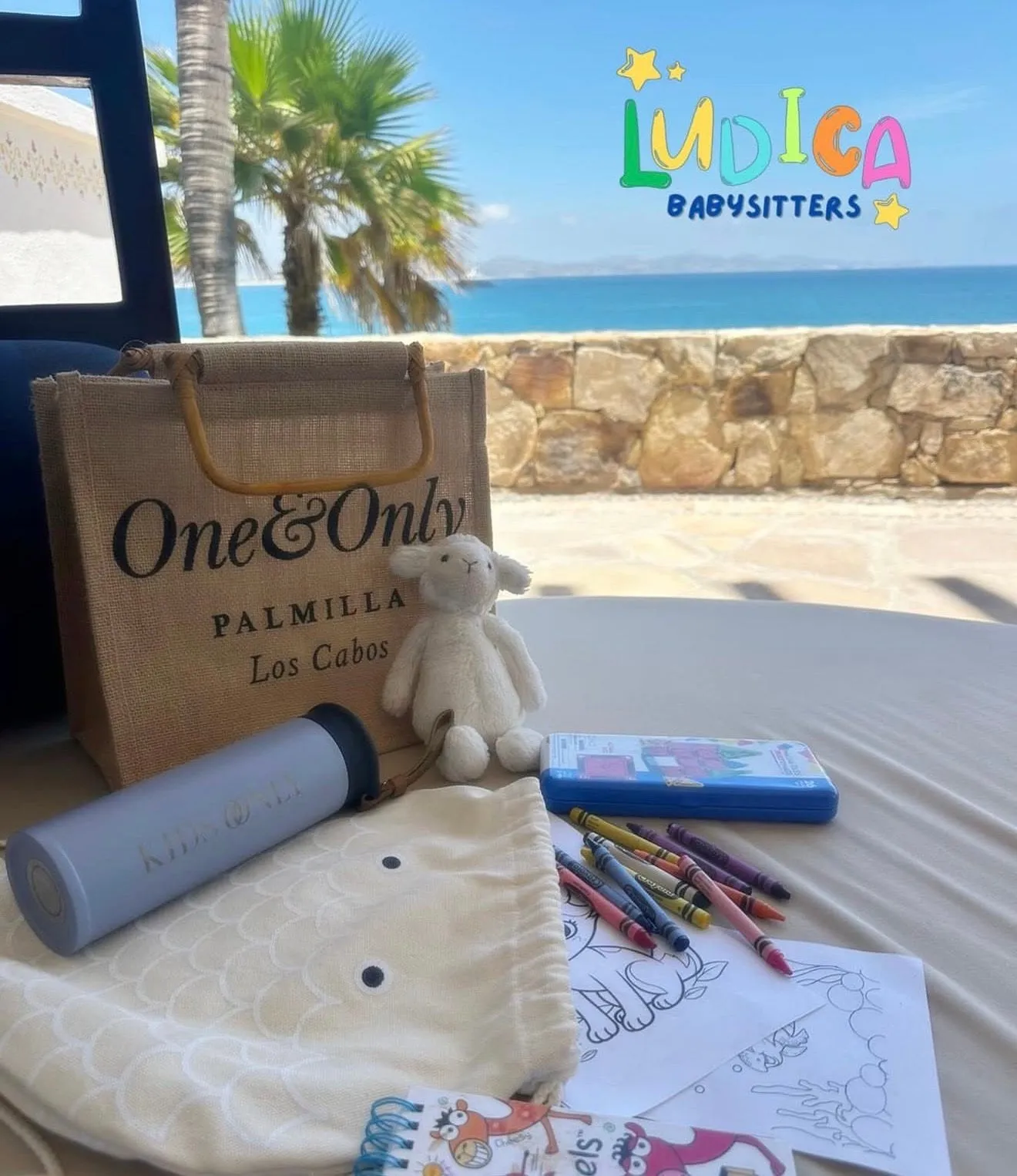 A welcome kit laid out for a family — tote, soft toy, blanket, crayons and a colouring book, the sea behind