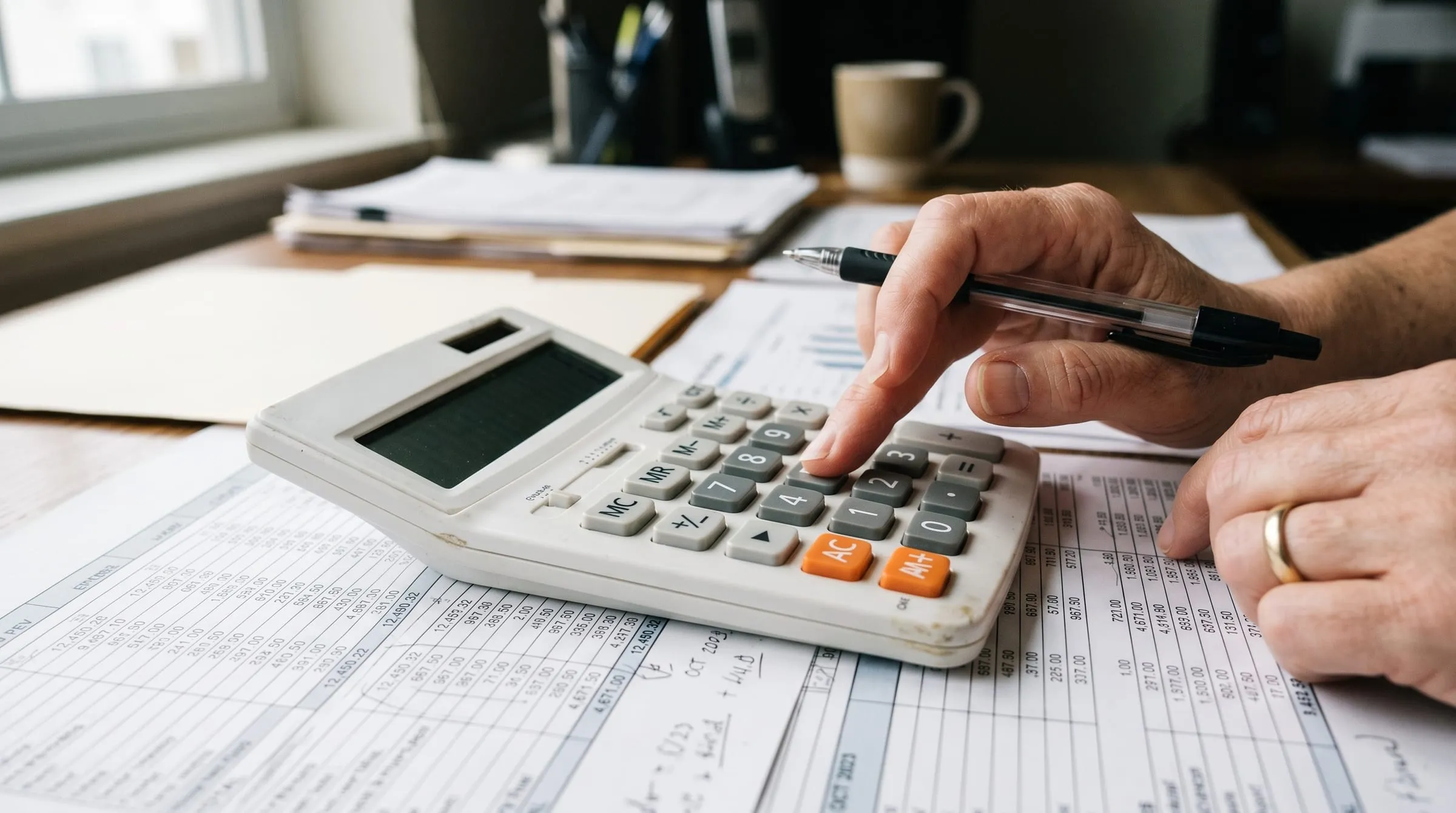 A hand working through a column of figures on a calculator, ledgers spread across the desk