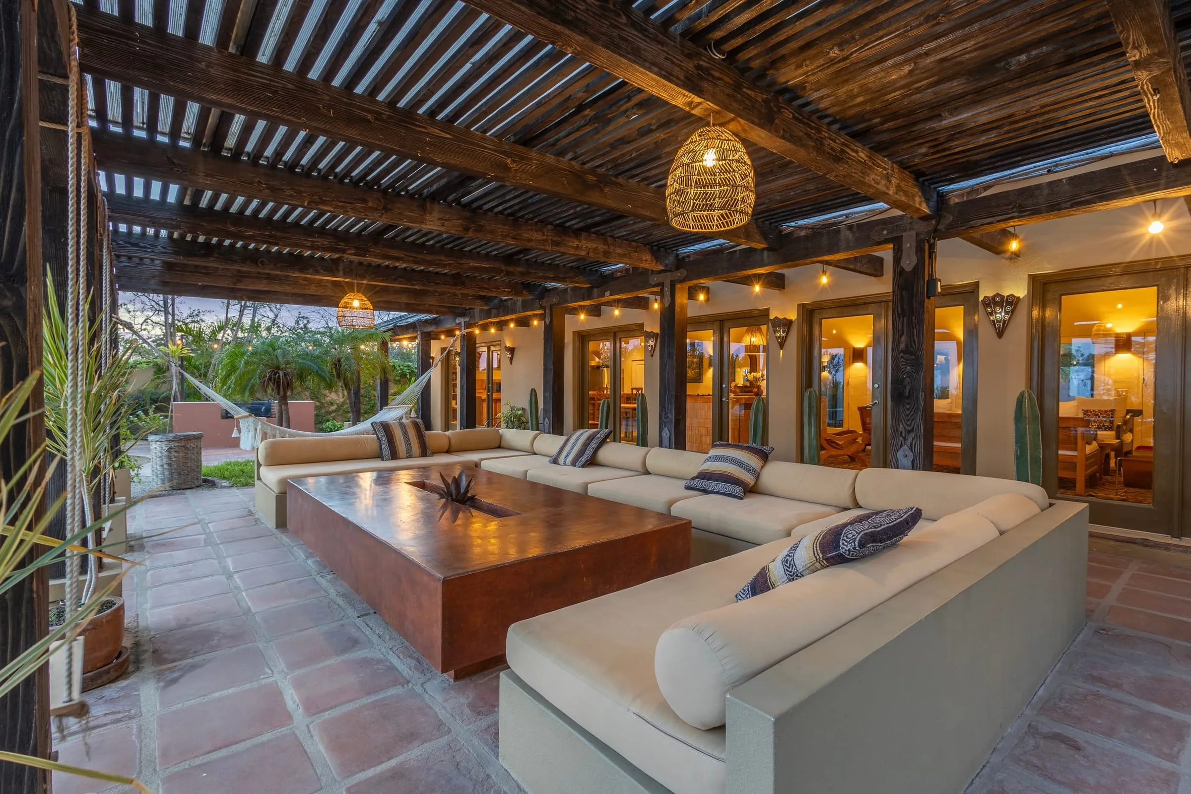 The covered terrace under its pergola, sofas around a long table, lit at dusk